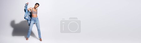 Foto de Modelo afroamericano con pelo corto y pendientes dorados posando en chaqueta vaquera, top con estampado animal y jeans sobre fondo gris, concepto de moda vaquera, banner - Imagen libre de derechos
