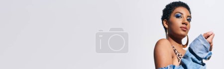 Foto de Vista de bajo ángulo del modelo afroamericano joven de moda con pendientes dorados mirando a la cámara y posando en chaqueta de mezclilla mientras está de pie aislado en gris, concepto de moda de mezclilla, pancarta - Imagen libre de derechos