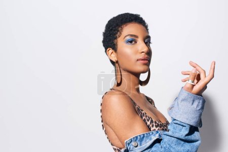 Trendy african american woman with short hair and vivid eyeshadow posing in top with animal print and denim jacket while standing on grey background, denim fashion concept