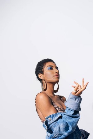 Téléchargez les photos : Jeune femme afro-américaine aux cheveux courts avec un maquillage audacieux posant en haut avec imprimé animal et veste en denim tout en regardant loin isolé sur gris, concept de mode en denim - en image libre de droit