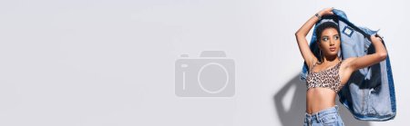 Téléchargez les photos : Jeune et jeune modèle afro-américain aux cheveux courts avec un maquillage vif tenant une veste en denim tout en posant en haut avec un imprimé léopard et un jean sur fond gris, concept de mode en denim, bannière - en image libre de droit