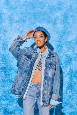 Cheerful and young african american woman with bold makeup touching beret while posing in denim jacket and jeans on blue textured background, stylish denim attire