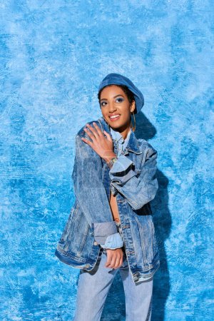 Jeune femme afro-américaine souriante avec un maquillage vif et un béret posant en jeans et veste en denim tout en se tenant debout sur un fond texturé bleu, tenue en denim élégante