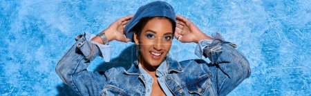 Retrato de modelo afroamericano sonriente y de pelo corto tocando la boina mientras posa en chaqueta de mezclilla y de pie sobre fondo azul texturizado, elegante atuendo de mezclilla, pancarta 