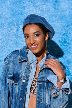 Cheerful african american model in beret, golden earrings and denim clothes looking at camera while standing near blue textured background, stylish denim attire