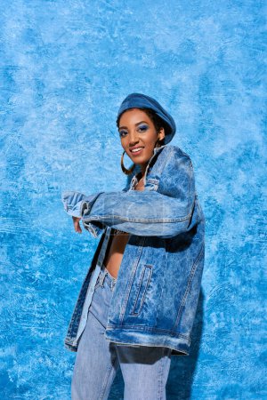 Fröhliches junges afrikanisch-amerikanisches Model mit lebhaftem Make-up, das Baskenmütze, Jeans und Jeansjacke trägt, während es auf blauem strukturiertem Hintergrund posiert, stylische Jeans-Kleidung