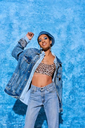 Positive african american model with bold makeup wearing beret, top with animal print and denim jacket while posing and standing near blue textured background, stylish denim attire