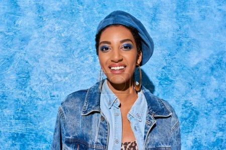 Retrato de mujer afroamericana joven y positiva con maquillaje audaz en boina y chaqueta de mezclilla mirando a la cámara sobre fondo azul texturizado, elegante atuendo de mezclilla