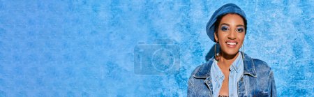 Cheerful african american model with bold eyeshadow and golden earrings wearing beret and denim jacket while standing on blue textured background, stylish denim attire, banner 