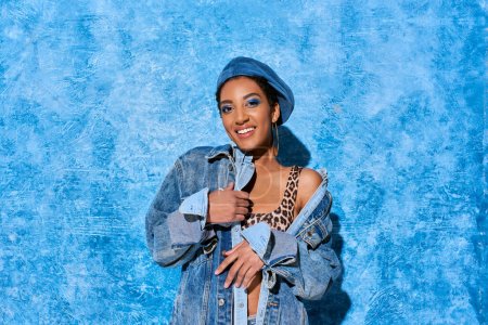 Téléchargez les photos : Femme afro-américaine à la mode et souriante avec un maquillage audacieux et un béret posant en haut avec une impression animale et une veste en denim sur fond texturé bleu, une tenue en denim élégante - en image libre de droit