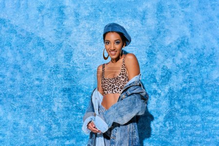 Fashionable african american woman in beret and top with animal print smiling at camera and posing in denim jacket on blue textured background, stylish denim attire