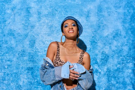 Positive and young african american woman with bold makeup and golden earrings posing in beret and denim jacket on blue textured background, stylish denim attire