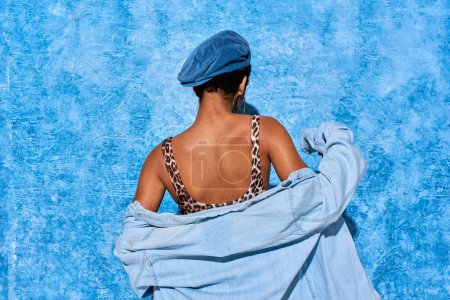 Rückenansicht einer kurzhaarigen und trendigen afrikanisch-amerikanischen Frau in Baskenmütze, Oberteil mit Animal Print und Jeanshemd auf blau strukturiertem Hintergrund, stylische Jeans-Kleidung