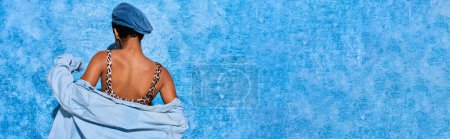 Vista posterior de la joven afroamericana posando en boina, parte superior con estampado de animales y camisa de mezclilla mientras está de pie y posando sobre fondo azul texturizado, elegante atuendo de mezclilla, pancarta 