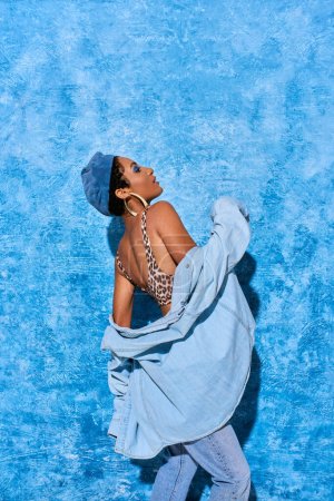 Foto de Vista lateral de la mujer afroamericana de moda en boina, parte superior con estampado de animales y camisa de mezclilla posando y de pie sobre fondo azul texturizado, elegante atuendo de mezclilla - Imagen libre de derechos