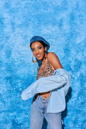 Modisches afrikanisch-amerikanisches Model mit goldenen Ohrringen, Baskenmütze und Jeanshemd, das in die Kamera lächelt, während es posiert und auf blauem strukturiertem Hintergrund steht, stylische Jeanskleidung