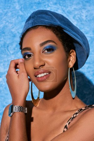 portrait de jeune femme afro-américaine avec un maquillage audacieux, béret et boucles d'oreilles dorées souriant à la caméra et posant sur fond bleu texturé, tenue en denim élégant