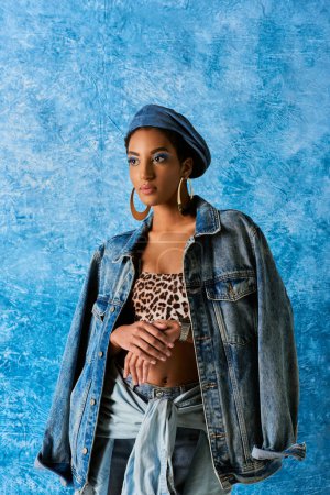 Modelo afroamericano de moda en boina y pendientes dorados posando en chaqueta de mezclilla y parte superior con patrón animal mientras está de pie sobre fondo azul texturizado, elegante atuendo de mezclilla