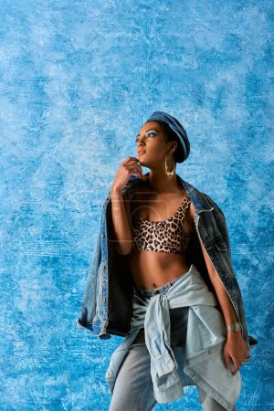 Modelo afroamericano de moda en boina y pendientes dorados mirando hacia otro lado mientras posan en la parte superior con patrón animal y chaqueta de mezclilla sobre fondo texturizado azul, elegante atuendo de mezclilla