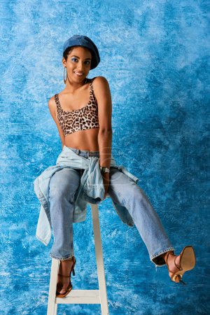 Volle Länge der fröhlichen afrikanisch-amerikanischen Frau in Baskenmütze, Top mit Animal Print und Jeans sitzt auf Stuhl und blickt in die Kamera auf blauem strukturierten Hintergrund, stilvolle Jeans-Kleidung