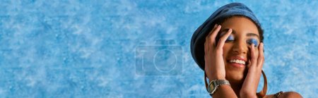 Trendy african american woman with bold makeup and denim beret smiling and touching face on blue textured background with copy space, stylish denim attire, banner 