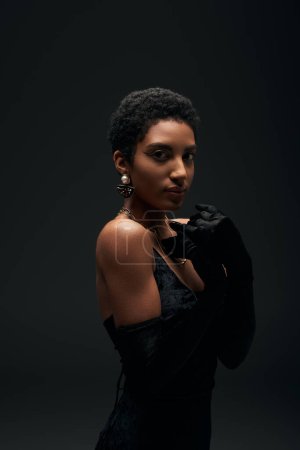 Elegant short haired african american woman with evening makeup posing in dress, gloves and golden accessories isolated on black, high fashion and evening look