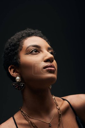 Retrato de mujer afroamericana elegante y de pelo corto con maquillaje de noche y accesorios dorados mirando hacia otro lado y de pie aislado en negro, alta moda y aspecto de noche