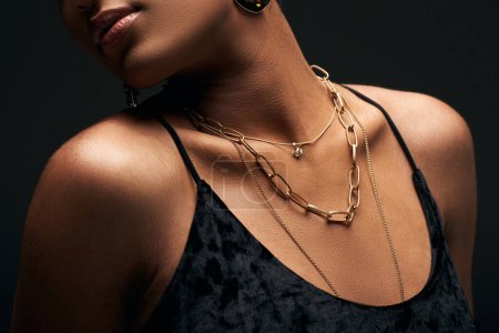 Cropped view of fashionable african american woman in evening dress and with golden necklaces posing isolated on black with lighting, high fashion and evening look, close up, details 