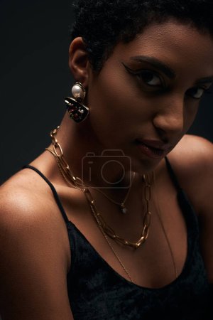 Retrato de modelo afroamericano de moda con maquillaje de noche y accesorios dorados mirando a la cámara aislada en negro con iluminación, alta moda y aspecto de noche