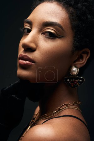 Vista de cerca de la joven mujer afroamericana segura y elegante con maquillaje de noche y accesorios dorados mirando a la cámara aislada en negro, alta moda y aspecto de noche
