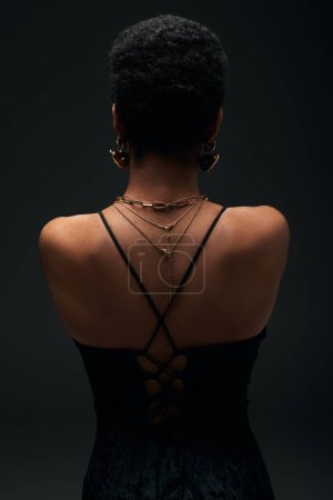Vista posterior de la mujer afroamericana de moda con pelo corto y accesorios de oro de pie aislado en negro con iluminación, alta moda y aspecto de noche, joyas, femenino, sensualidad 
