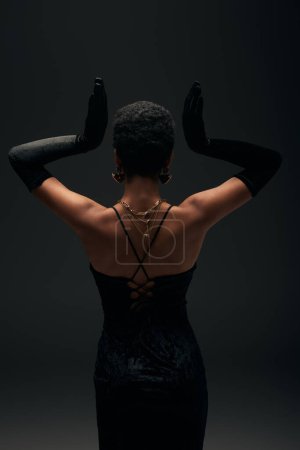 Back view of elegant african american woman in evening dress, gloves and golden accessories posing isolated on black with lighting, high fashion and evening look, feminine sensuality 