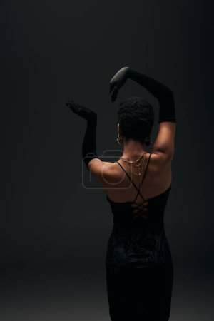 Téléchargez les photos : Vue arrière de l'élégante femme afro-américaine aux cheveux courts en gants, robe de soirée et accessoires dorés posant isolée sur look noir, haute couture et soirée - en image libre de droit