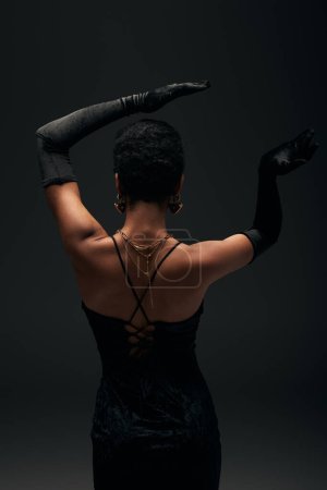 Rückansicht einer modischen afrikanisch-amerikanischen Frau in Handschuhen, goldenen Accessoires und Abendkleid, die isoliert auf schwarzem High Fashion und Abendlook posiert