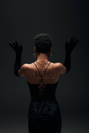 Rückenansicht des trendigen afrikanisch-amerikanischen Modells mit goldenen Accessoires und sortiertem Haar posiert in Handschuhen und Abendkleid isoliert auf schwarz, High Fashion und Abendlook