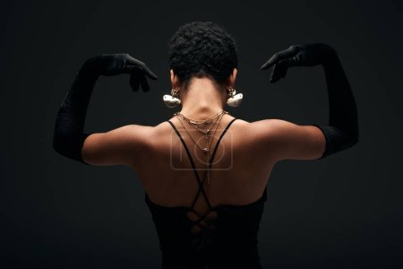 Back view of elegant african american woman in gloves and golden accessories posing in light while standing isolated on black, high fashion and evening look