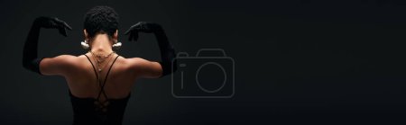Back view of fashionable young african american woman in gloves and golden accessories posing in lighting while standing isolated on black, high fashion and evening look, banner 