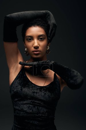 Photo for Portrait of elegant african american model in evening dress, golden accessories and gloves posing and looking at camera while standing isolated on black, high fashion and evening look - Royalty Free Image