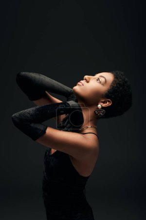Foto de Vista lateral del modelo afroamericano de moda en guantes elegantes y vestido mirando hacia arriba mientras está de pie bajo la iluminación aislada en negro, alta moda y aspecto de noche - Imagen libre de derechos