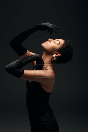 Téléchargez les photos : Vue latérale de l'élégante femme afro-américaine aux cheveux courts en gants, robe et accessoires dorés posant avec les yeux fermés isolés sur le noir, look haute couture et soirée - en image libre de droit