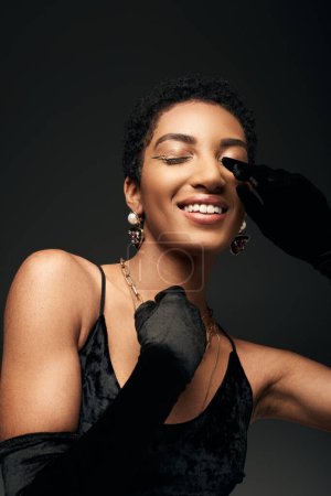 Portrait of fashionable and smiling african american woman in dress and gloves touching face while closing eyes and standing isolated on black, high fashion and evening look