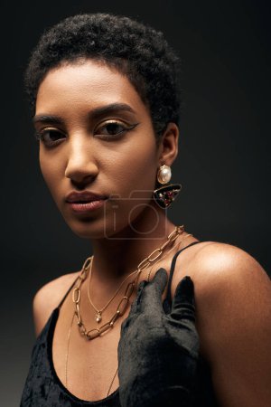 Jeune femme afro-américaine aux cheveux courts avec maquillage du soir et accessoires dorés posant en robe et gant isolé sur look noir, haute couture et soirée