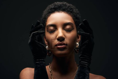 Retrato de mujer afroamericana elegante y de pelo corto con maquillaje de noche, cadenas doradas, pendientes y guantes posando con los ojos cerrados aislados en negro, alta moda y aspecto de noche
