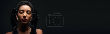 Elegante und kurzhaarige afrikanisch-amerikanische Frau mit Abendschminke, Accessoires und Handschuhen, die mit geschlossenen Augen isoliert auf schwarzem High Fashion und Abendlook steht, Banner 