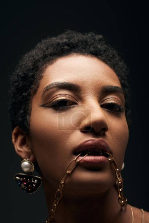 Retrato de modelo afroamericano de pelo corto y elegante con maquillaje de noche que sostiene el collar de oro en la boca y mirando a la cámara aislada en negro, alta moda y aspecto de noche
