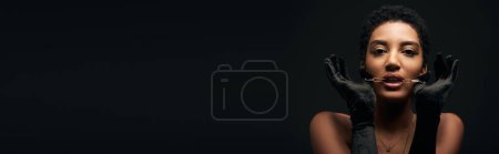 Foto de Modelo afroamericano de moda con maquillaje de noche que usa guantes y sostiene la cadena dorada cerca de la boca aislada en negro con iluminación, alta moda y apariencia nocturna, pancarta - Imagen libre de derechos