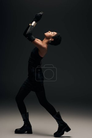 Photo for Side view of fashionable african american model in little dress, gloves and boots posing while standing in lighting on black background, high fashion and evening look - Royalty Free Image