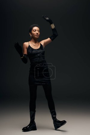 Elegantes afrikanisch-amerikanisches Model in kleinem Kleid, Handschuhen und goldenen Accessoires mit geschlossenen Augen auf schwarzem Hintergrund, High Fashion und Abendlook