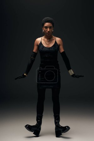 Full length of short haired african american woman in little dress, gloves and boots looking at camera while standing on black background, high fashion and evening look