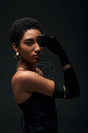 Modisches und kurzhaariges afrikanisch-amerikanisches Model in Kleid, Accessoires und Handschuh bedeckendem Auge, während es isoliert auf schwarzem High Fashion und Abendlook posiert